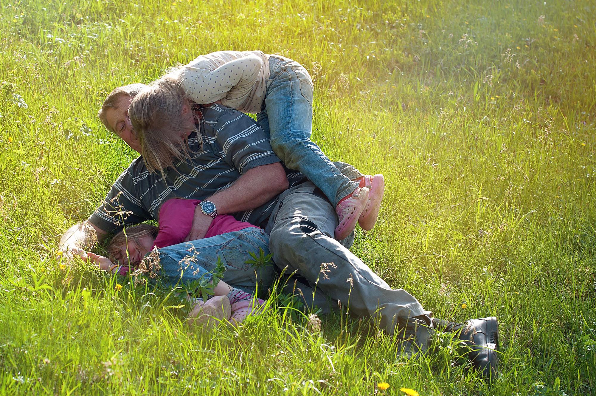 Dad with two little girls obviously enjoying fun family time with dad sitting on the grass rolled on one side, while one of the little girls climbed on top of him, while the other little girl laughs rolled in the grass next to her dad. 