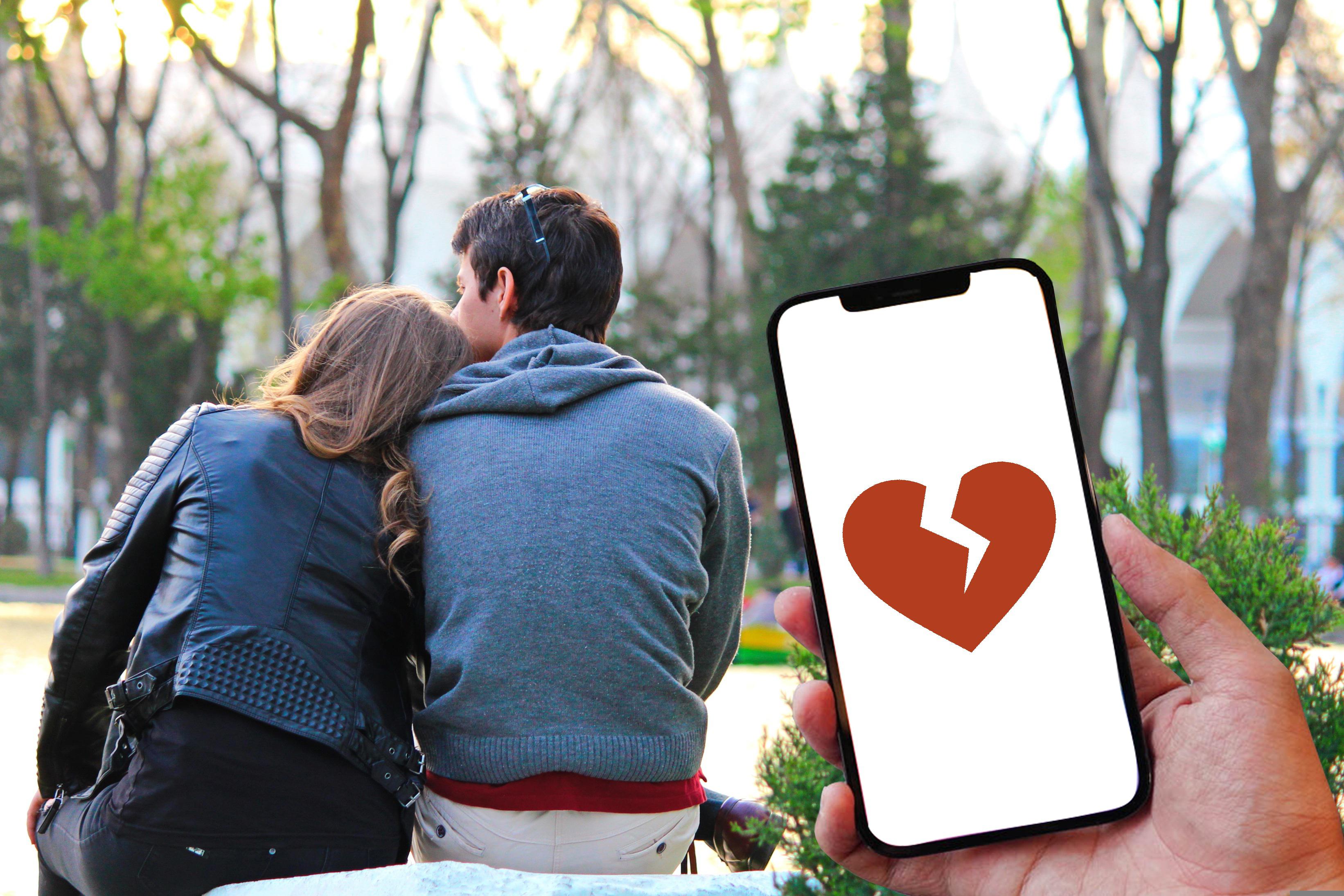 In-love couple, female's head leaning against male's shoulder, sitting on a bench in the park, with a phone picturing a broken heart in the forefront. 