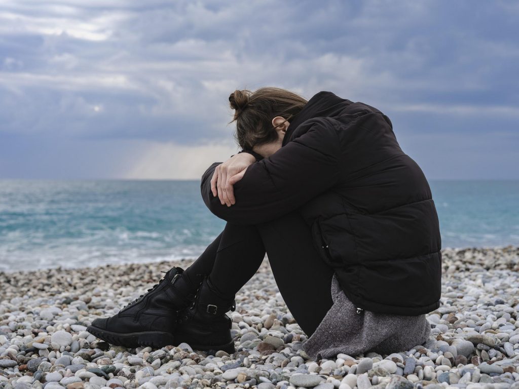 Woman sitting on the beach by the ocean, visibly upset, head down on her knees, obviously worried and upset. 